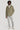 Sage-colored field jacket with chest pockets and hood, worn with white shirt, light gray pants, and white sneakers.
